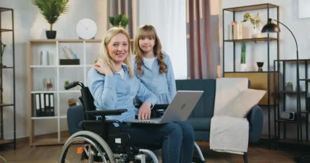 Attrayant heureux souriant femme handicapée aimante en fauteuil roulant regardant la caméra avec sa jolie fille positive de 10 ans dans le salon joliment décoré — Video