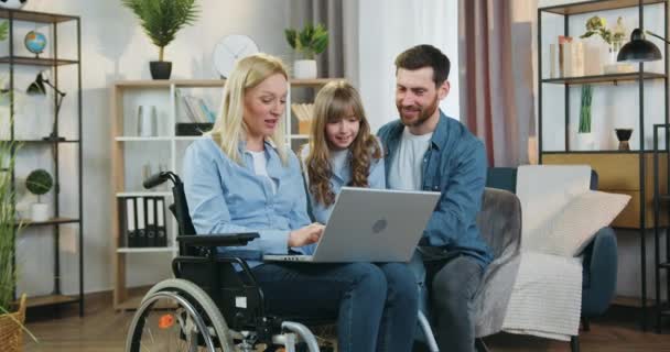 Idylle familiale de femme handicapée souriante adorable en fauteuil roulant utilise ordinateur portable avec la fille mignonne de 10 ans et mari barbu aimant joyeux à la maison — Video