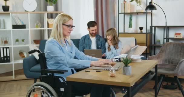 Mujer con discapacidad positiva de buen aspecto en silla de ruedas que trabaja en el ordenador portátil en casa, mientras que su marido barbudo cuidado guapo enseñar a su linda hija de 10 años de edad cómo utilizar el ordenador portátil — Vídeo de stock