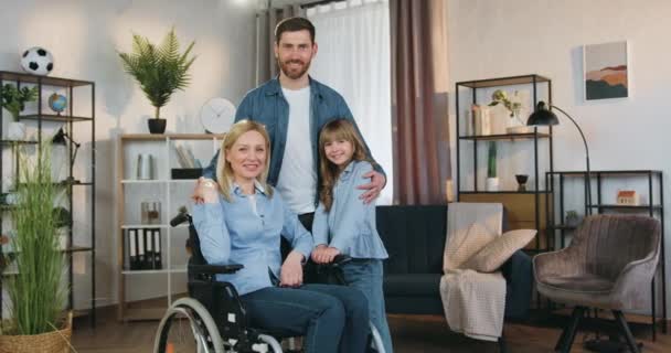 Retrato familiar de la hermosa sonriente mujer discapacitada de pelo claro feliz en silla de ruedas que mira a la cámara junto con su cariñoso esposo barbudo y su encantadora hija alegre en casa — Vídeo de stock