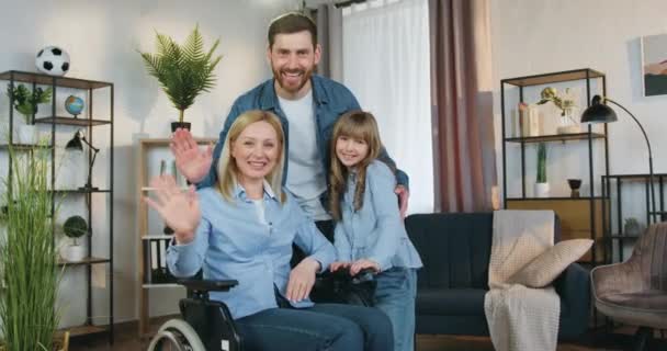 Jolie femme handicapée affectueuse et joyeuse en fauteuil roulant regardant la caméra avec son mari barbu bienveillant et sa jolie fille de 10 ans et ils saluent tous les mains — Video