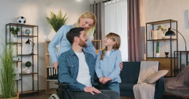 Attrayant aimant sourire barbu homme handicapé en fauteuil roulant posant sur la caméra avec sa fille mignonne de 10 ans et assez heureuse épouse joyeuse dans le salon confortable — Video