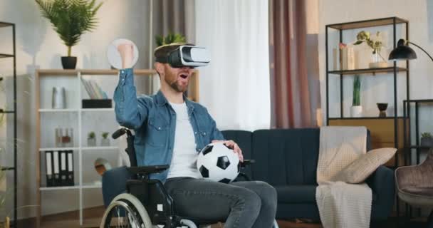 Attraktiv glad glad skäggig funktionshindrade man i rullstol efter olycka titta på fotbollsmatch med hjälp av virtuell verklighet headset i vackert inredda vardagsrum — Stockvideo