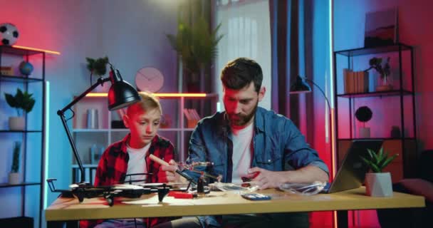 Привлекательный концентрированный бородатый отец паяльник важных деталей под лампой дома в вечернее время и его заинтересованный 10-летний сын наблюдает за работой отцов — стоковое видео