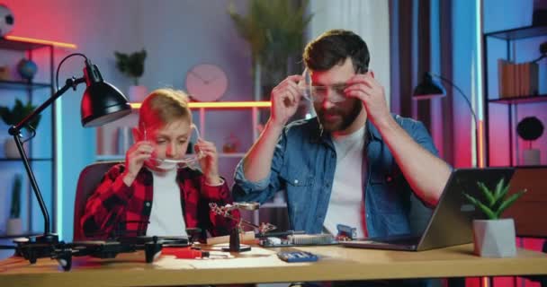 Concept de connaissance quotidienne humaine où sympathique confiant adolescent positif garçon et son père portant des lunettes de protection avant soudure détails sur l'électronique à la maison le soir — Video