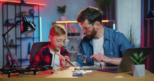 Knappe nieuwsgierige tiener jongen in een beschermende bril kijken door vergrootglas hoe zijn vaardige bebaarde vader hechten kleine onderdelen op elektronica met behulp van soldeerbout thuis in de avond — Stockvideo