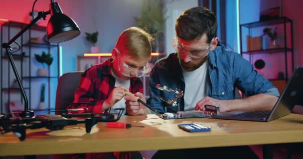 Guapo cuidado hábil padre barbudo adulto y su curioso hijo attantive tanto en gafas protectoras soldando pequeños detalles en el panel electrónico juntos sosteniendo soldador — Vídeos de Stock