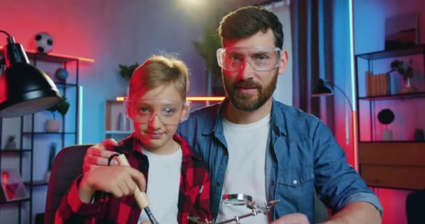 Primer plano de guapo padre barbudo adulto alegre y lindo adolescente en gafas protectoras que mirando a la cámara con expresión de cara feliz durante la soldadura en casa por la noche — Vídeo de stock