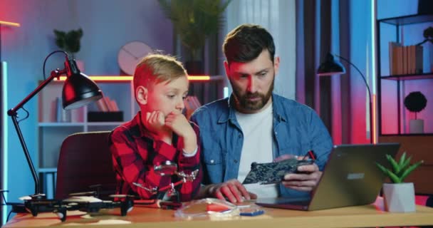 Привлекательный опытный взрослый бородатый отец разговаривает со своим любопытным сыном-подростком об электронной панели с компонентами и читает информацию на компьютере дома вечером — стоковое видео