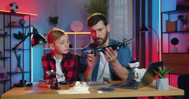 Portrait de beau père barbu habile attentionné confiant qui explique comment utiliser un drone à son fils adolescent curieux dans une chambre confortable avec des veilleuses — Video