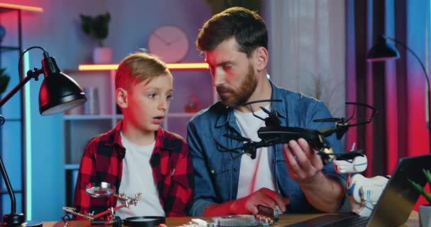 Knap zelfverzekerde moderne volwassen bebaarde vader zit aan tafel samen met zijn nieuwsgierige zoon en ze praten over drone in vaders handen, slow motion — Stockvideo