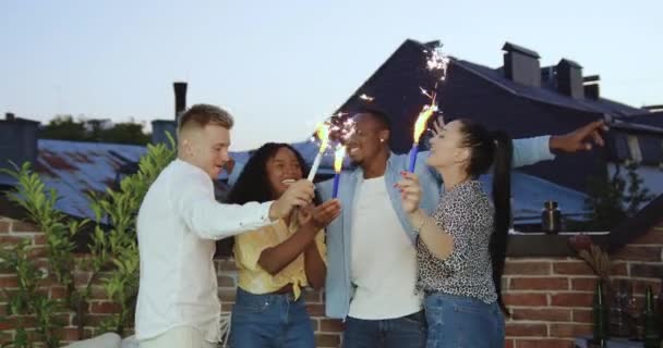 Gutmütig unbeschwert lächelnd fröhliche junge Mischlingsfreunde feiern Sommerfest auf der Terrasse mit funkelnden Lichtern bei Sonnenuntergang — Stockvideo