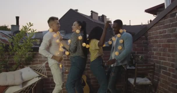 Atractivo positivo de buen humor jóvenes amigos diversos que celebran la fiesta de verano con bombillas amarillas, bailando en la terraza por la noche, vista frontal — Vídeo de stock