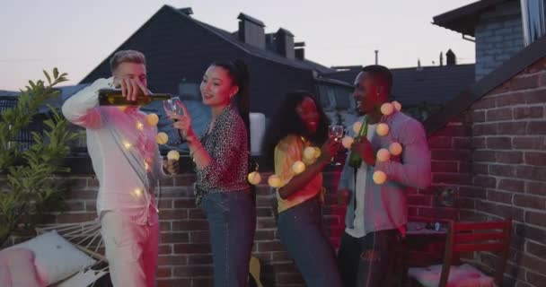 Schöne gemischte Rasse zufrieden zwei Mädchen und die gleichen zwei gutaussehenden Jungs tanzen mit Gläsern Wein während Freunde Party auf dem großen Balkon mit kleinen runden Lampen bei Sonnenuntergang — Stockvideo