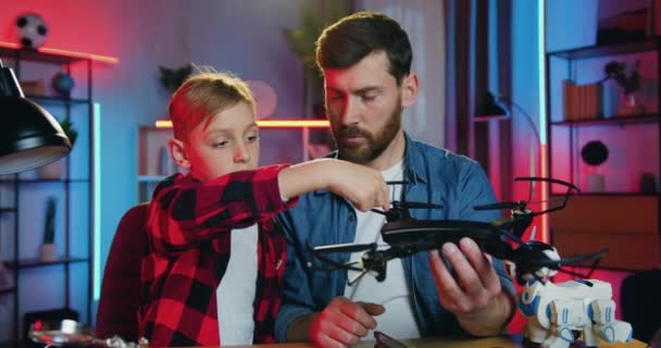 Attractive positive skillful bearded father and smiling 12-aged boy sitting together at the table in cozy room in the evening and talking about modern drone in fathers hands — Stockvideo
