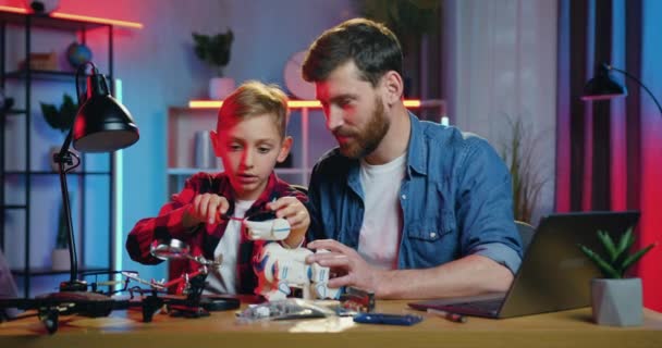Handsome happy smiling bearded father and his curious son repairing robot using screwdriwers sitting at workplace in the evening,front view — стоковое видео