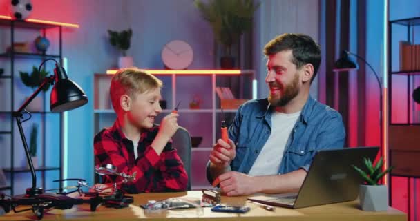 Good-looking cheerful adult bearded man and his joyful smiling teen son playing with screwdriwers like swords during repairing electronic panel at home in the evening — Video