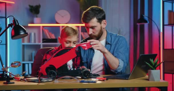Handsome happy caring adult bearded father rejoycing together with his curious 12-aged son after they successfully repairing car toy in cozy room in the evening — стоковое видео