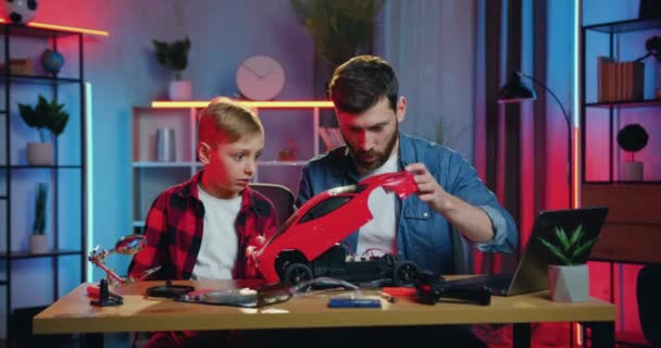 Good-looking surprised adult bearded man taking off toy cars top and studied the design of inside car together with his curious teen son at home with night lightings effects — стоковое видео