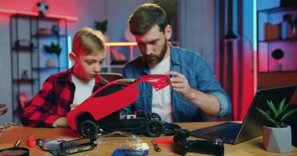 Good-looking positive skilled adult bearded man taking off toy cars top and together with his interested teen son studied the cars content at home — Αρχείο Βίντεο