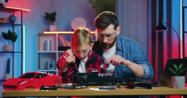 Portrait of attractive serious adult bearded man and teen boy both in protective glasses soldering toy cars wires or details using soldering iron at home in the evening — Αρχείο Βίντεο