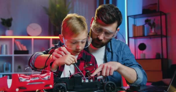 Good-looking satisfied loving adult bearded father in protective glasses teaching to solder small details of toy car his curious smart teen son in protective glasses too — Vídeo de Stock