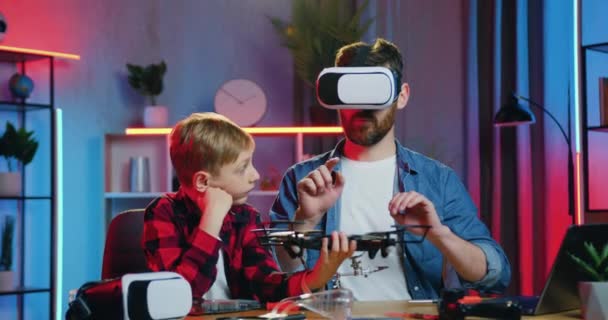 Handsome positive teen boy holding modern flying aparat in his hand while confident bearded man studied its construction using augmented reality goggles — Αρχείο Βίντεο