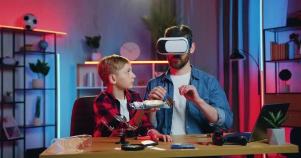 Handsome confident high-skilled adult bearded man in augmented reality headset studying electronic panel which is held by his positive teen son,close up,slow motion — Vídeo de Stock