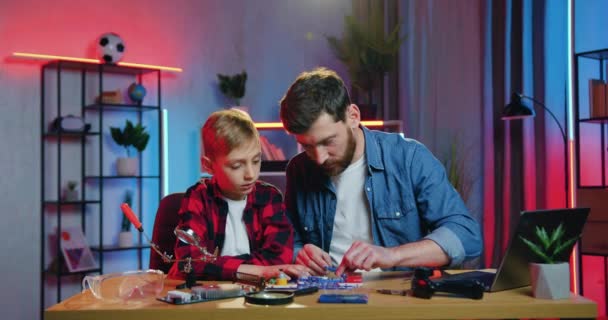 Handsome caring confident positive bearded father helping to assemble the small parts of constructor together with his nice interested teen son at home — Wideo stockowe