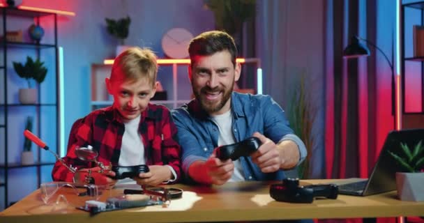 Концепция семьи отдыха, где привлекательный счастливый улыбающийся бородатый папа и сын-подросток наслаждаются видеоиграми и празднуют победу с поднятыми руками, вид спереди — стоковое видео
