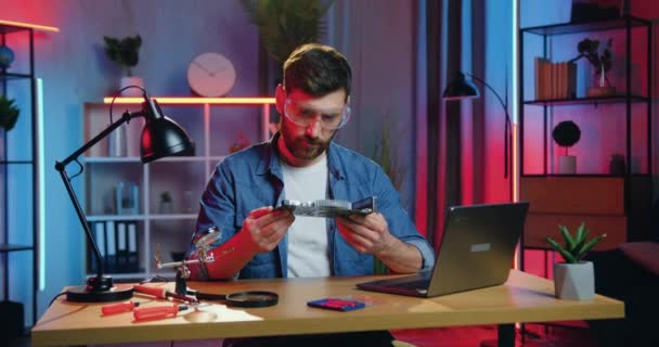 Atraktivní vážné sebevědomí zkušený vousatý muž v ochranných brýlích s ohledem na počítačové desce v rukou a čtení návod na jeho strukturu na obrazovce notebooku — Stock video