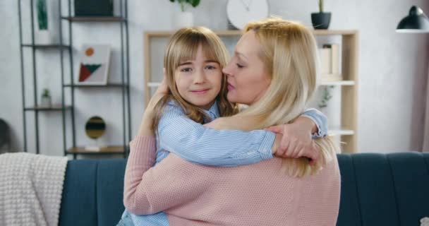 Concepto familiar de adorable madre de pelo claro y cariñosa que abraza y besa a su alegre hija de 12 años en una acogedora sala de estar, cámara lenta — Vídeo de stock