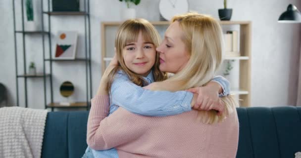 Concept familial d'une belle mère aux cheveux clairs et attentionnée qui étreint et embrasse sa joyeuse fille de 12 ans dans un salon confortable, au ralenti — Video