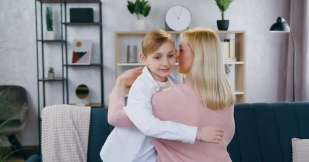 Portret van een knappe glimlachende tienjarige jongen die knuffelt met zijn liefdevolle 35-jarige moeder in prachtig ingerichte zitkamer — Stockvideo