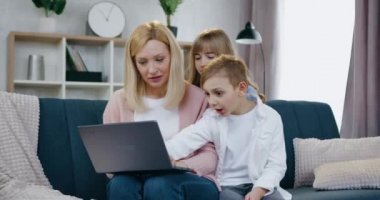 Güzel, mutlu, gülümseyen bir anne, akıllı, pozitif iki çocuğuyla birlikte evdeki yumuşak kanepede oturuyor ve heyecan verici videolar izlemek için dizüstü bilgisayar kullanıyor.