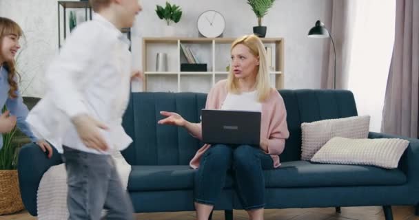 Attractive cheerful noisy playful boy and girl running around the couch on what sits their concentrated worried busy mother and working on laptop — Stock Video