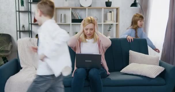 Problemen in afgelegen huis werk waar aantrekkelijke nerveuze drukke vrouw sloot haar oren zitten op de bank tijdens haar werk op de laptop om niet te horen het lawaai en geschreeuw van haar speelse kinderen rennen — Stockvideo