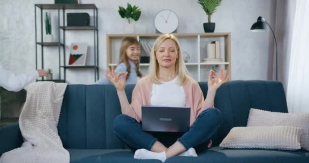 Atraente ativo alegre 12-aged irmão e irmã correndo ao redor do sofá em que sentado sua mãe muito calma de cabelos claros em pose de lótus com os olhos fechados e meditando — Vídeo de Stock