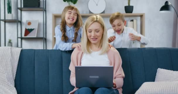 Bella felice madre sorridente che lavora sul computer portatile seduto sul divano e i suoi gioiosi bambini adolescenti giocosi che saltano fuori dietro il divano e la abbracciano — Video Stock