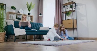 Mutlu, gülümseyen, açık renk saçlı bir annenin dizüstü bilgisayarda oturup oyun oynayan genç oğlu ve kızıyla yerde araba yarışı yaptığı bir aile.