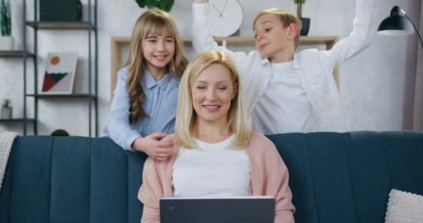 Urocza szczęśliwa uśmiechnięta 35-letnia matka siedząca z komputerem na kanapie i patrząca w kamerę z sympatyczną dwójką radosnych nastolatków, które przytulają ją za szyję — Wideo stockowe