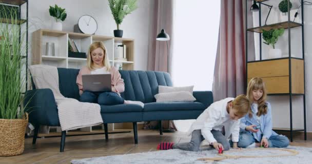 Familienfreizeitkonzept, wo eine lieblich fröhlich lächelnde, sorglose Mutter ihren Laptop benutzt, während ihre gut aussehenden, aktiven, verspielten zwei Kinder auf dem Fußboden mit einer Autoreise spielen — Stockvideo