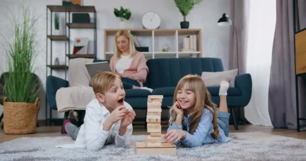 Schöne fröhliche fröhliche Teenager Bruder und Schwester haben Spaß zusammen beim Spielen mit Holzsteinen auf dem Boden und lachen, wenn Turm gebrochen ist, während ihre positiv fürsorgliche Mutter sie von — Stockvideo