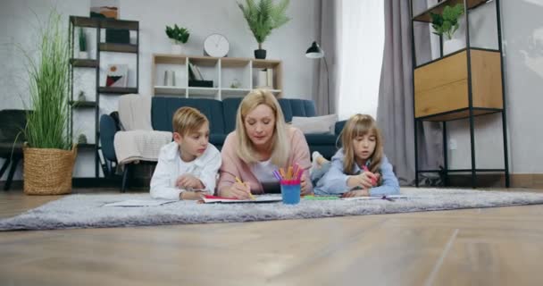 Családi szabadidő koncepció, ahol vonzó boldog szerető gondtalan anya fekszik a padlón a pozitív játékos gyerekekkel együtt, és rajzol velük — Stock videók
