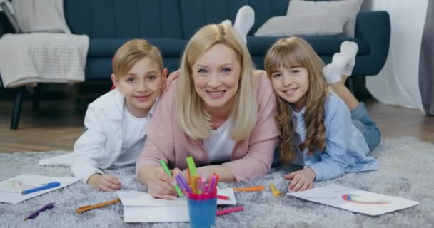 Ritratto di affascinante madre felice dai capelli chiari sorridente che guarda la macchina fotografica insieme al suo allegro figlio spensierato giocoso e figlia sdraiata sul pavimento durante il disegno congiunto — Video Stock