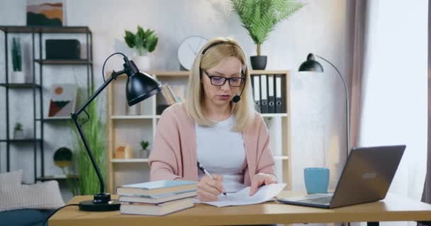 Mujer de negocios moderna inteligente bastante concentrada en auriculares sentados delante de la computadora portátil en la oficina acogedora en casa durante la reunión de vídeo con subordinados y discutir detalles del proyecto conjunto — Vídeos de Stock