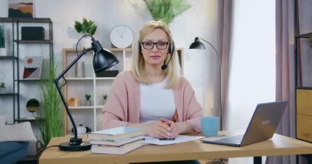 Привлекательная уверенная в хорошем настроении взрослая блондинка в наушниках и очках, смотрящая на камеру в домашнем офисе во время видеоконференции на компьютере с клиентами — стоковое видео