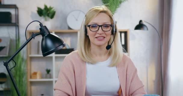 Piękna szczęśliwa uśmiechnięta blondynka w wieku 35 lat w słuchawkach w okularach patrząca w kamerę z zadowolonym wyrazem twarzy na tle przytulnego gabinetu — Wideo stockowe
