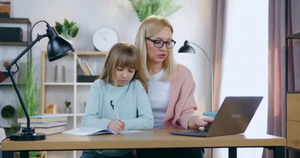 Vacker omtänksam kärleksfull 35-årig blond mamma i glasögon sitter framför datorn tillsammans med sin 12-åriga söta dotter och hjälpa henne med hemuppgiften — Stockvideo