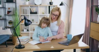 Çekici, mutlu, gözlüklü, gülümseyen, açık renk saçlı anne, akıllı kızına okul ödevinde yardım ediyor ve başarılı sonuçlardan keyif alıyor.